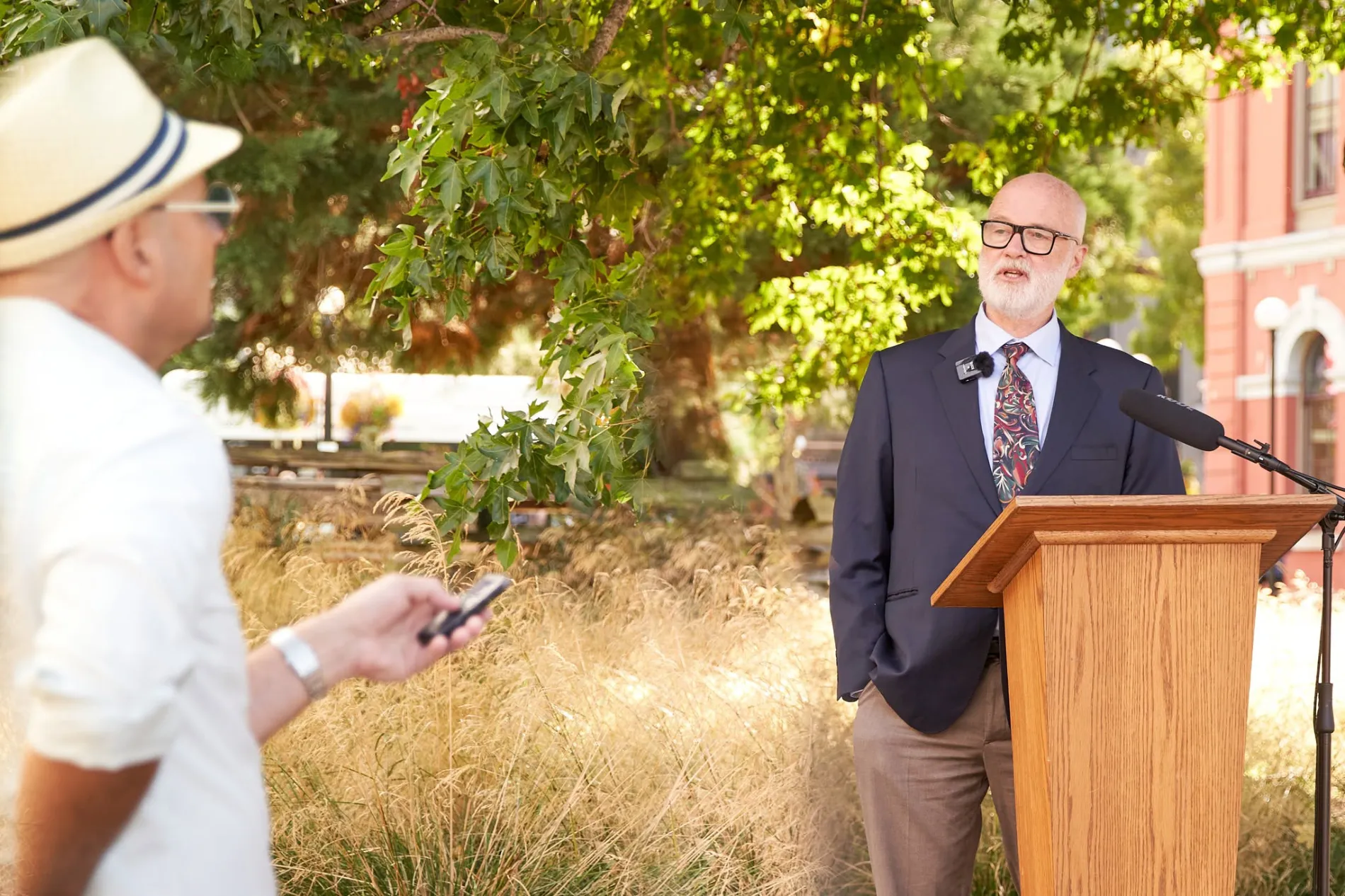 Bruce McGuigan speaking at a podium outdoors while a man in a straw hat holds out a phone to record