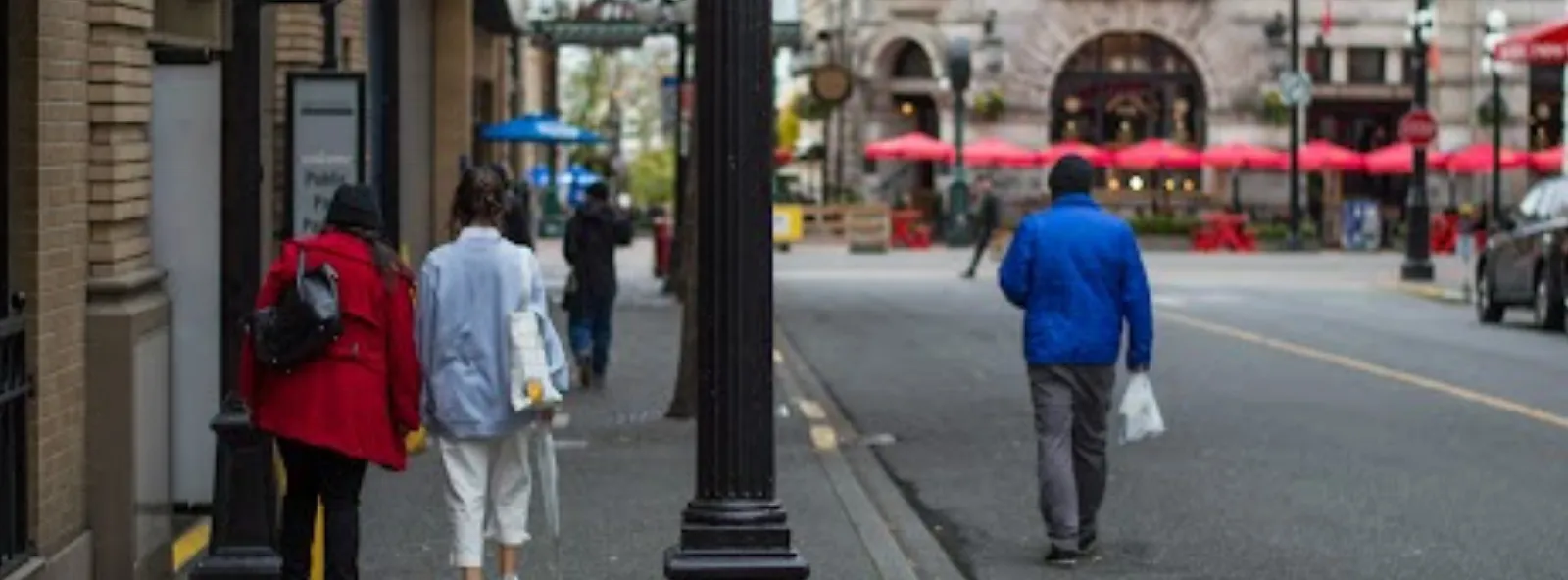 Pedestrians walk along a downtown Victoria street past a black lamp post, with red patio umbrellas in the background