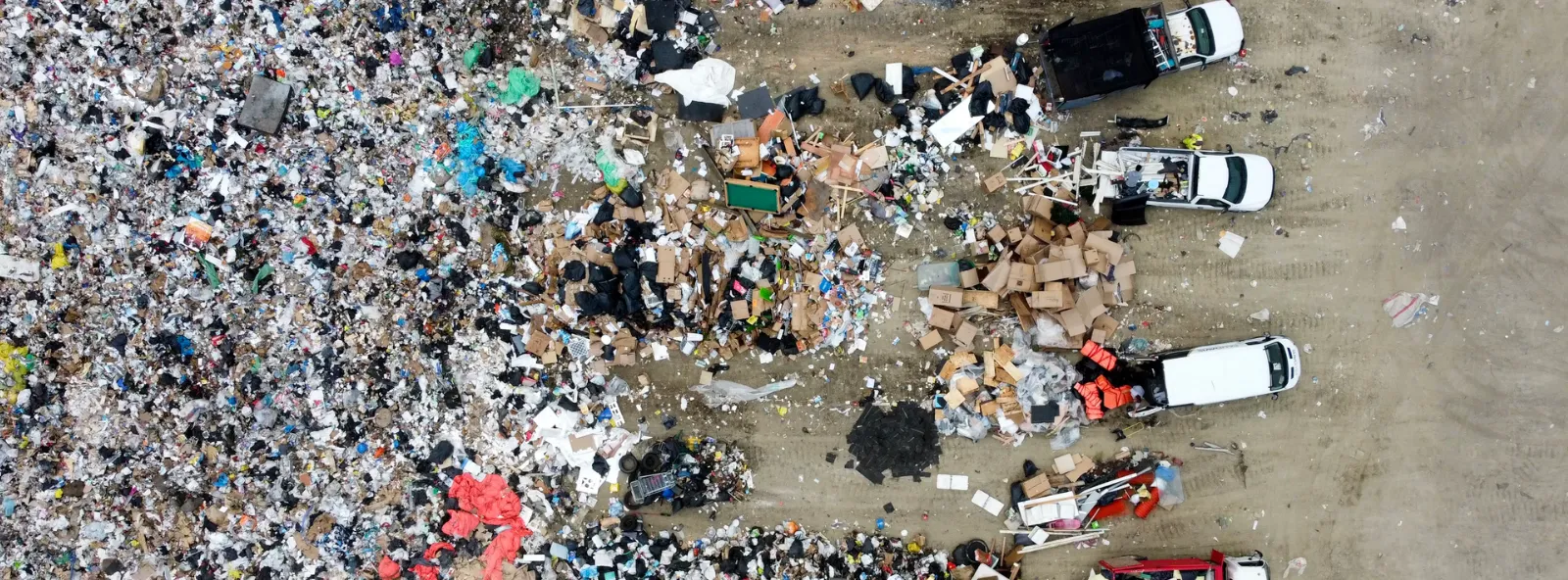 Aerial view of a landfill where trucks unload furniture, cardboard and household items beside a large expanse of mixed waste