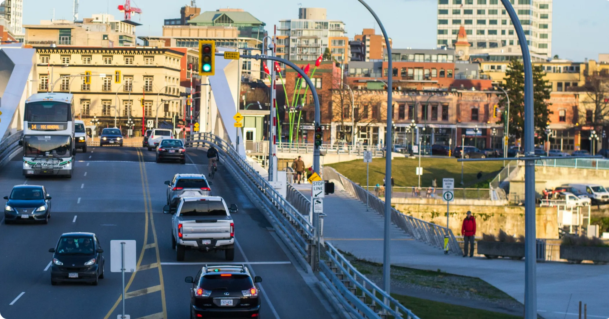 Cars, a BC Transit bus and a cyclist crossing the Johnson Street Bridge, with pedestrians on the walkway beside it.